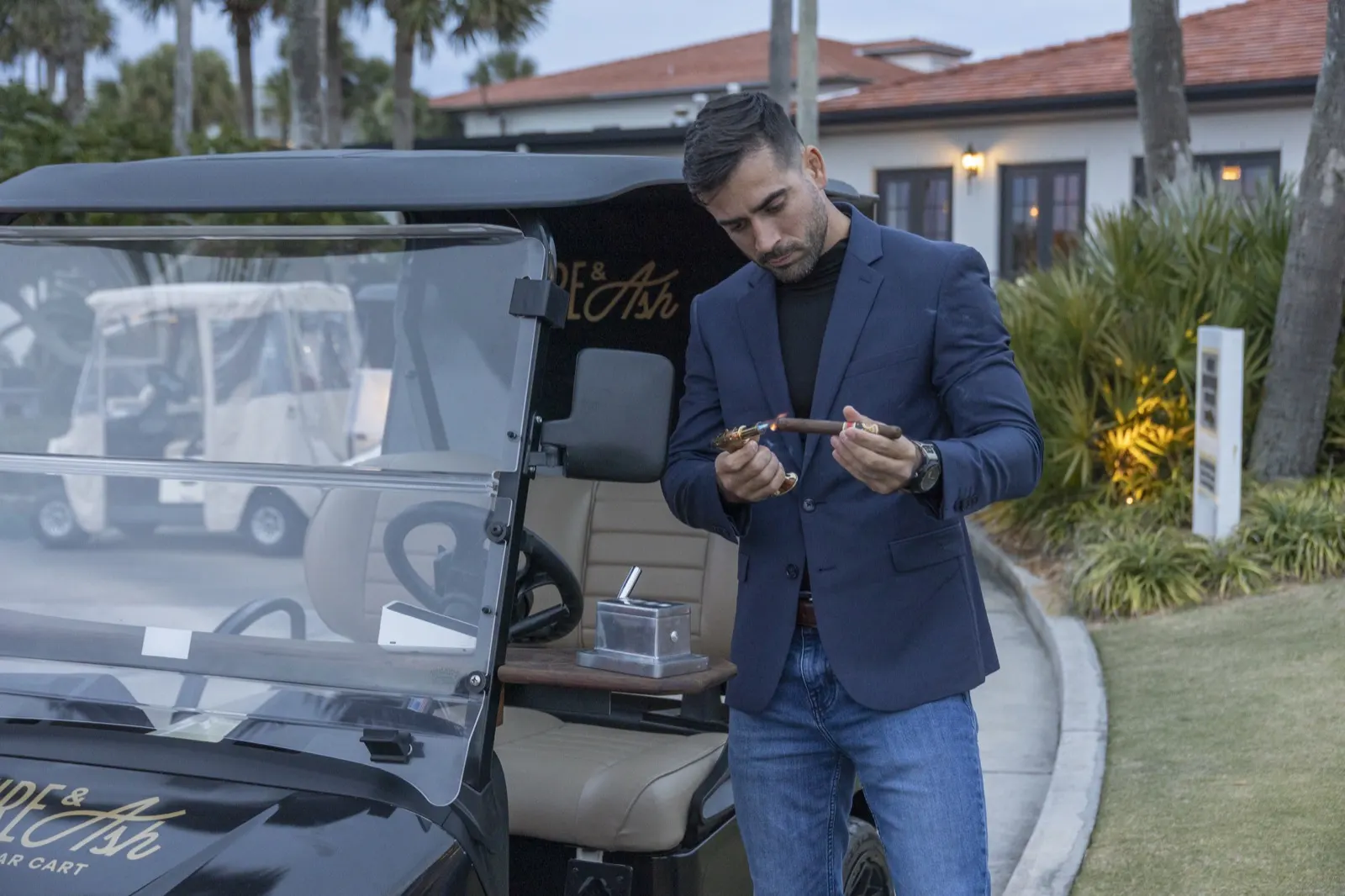 Carlos Lopez lighting a cigar beside the Fire and Ash golf cart at dusk