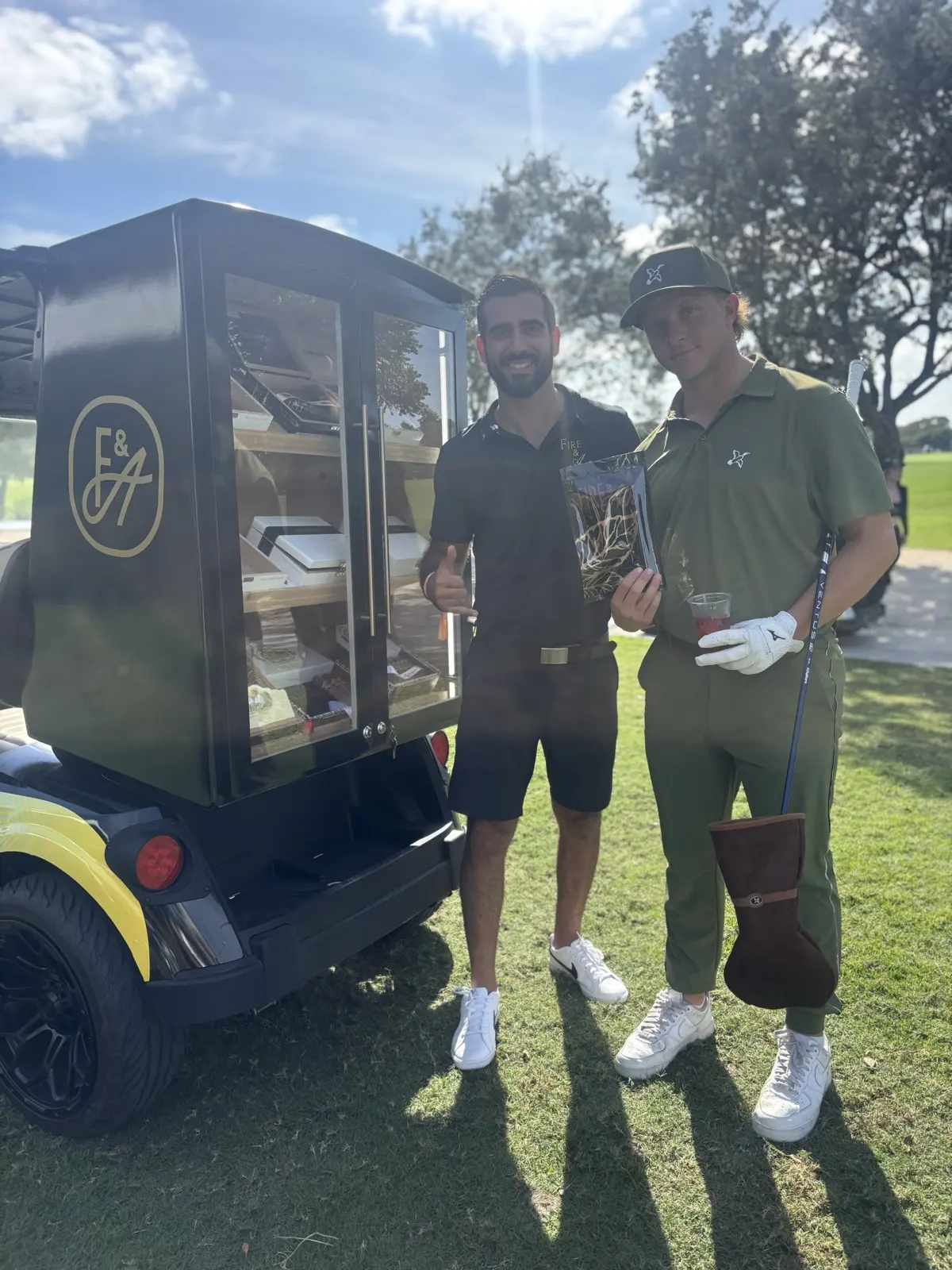 Two guests smiling beside the cigar golf cart humidor on the course