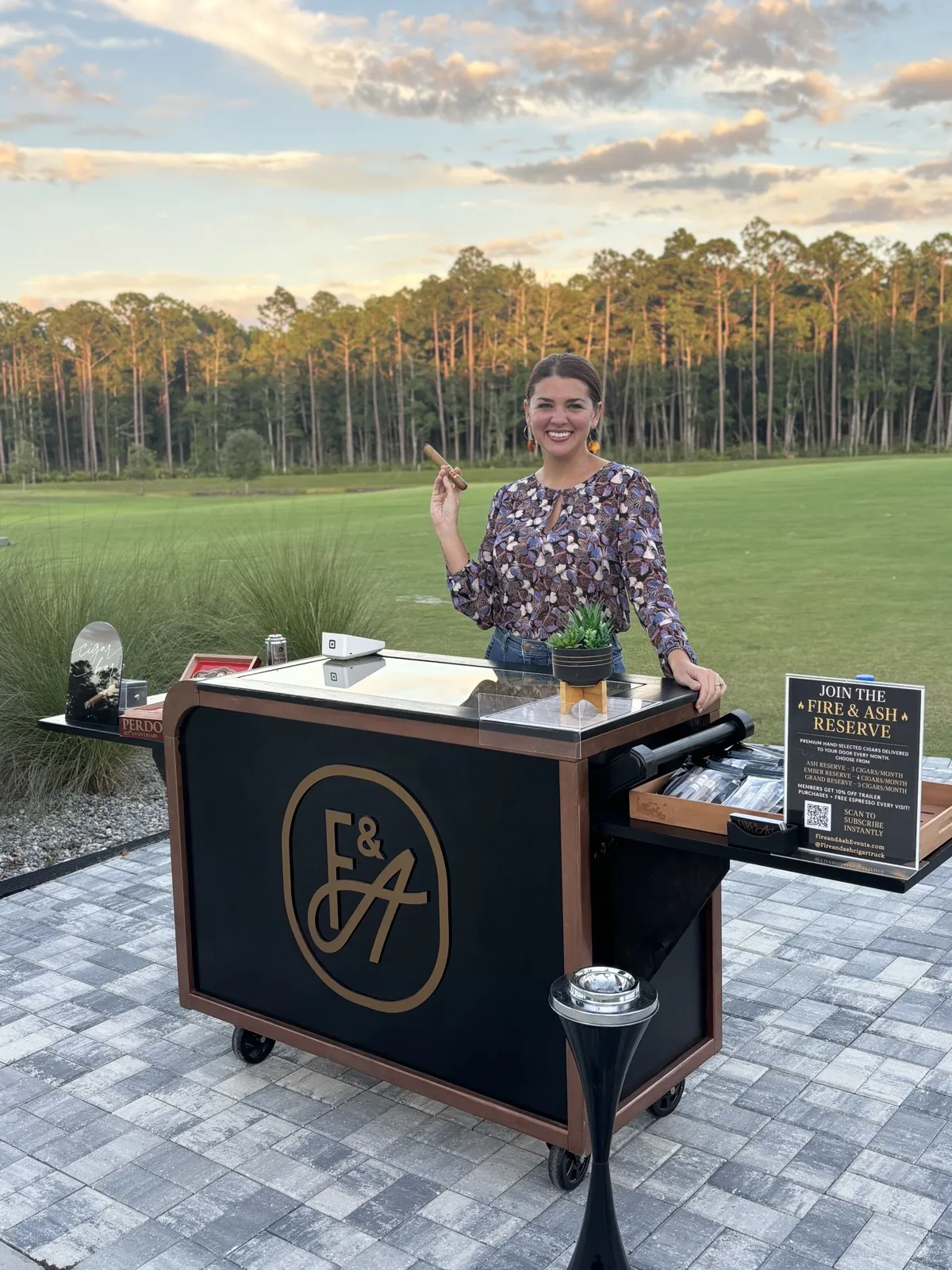 Hannah Lopez smiling behind the Fire and Ash cigar bar cart at a golf course event
